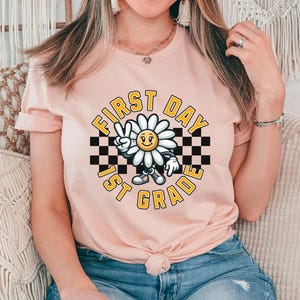 May include: A pink t-shirt with a graphic of a smiling daisy wearing a checkered headband and holding up a peace sign. The text "First Day 1st Grade" is written in a yellow circle around the daisy.