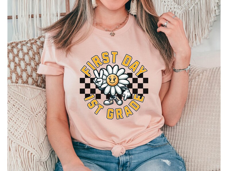 May include: A pink t-shirt with a graphic of a smiling daisy wearing a checkered headband and holding up a peace sign. The text "First Day 1st Grade" is written in a yellow circle around the daisy.