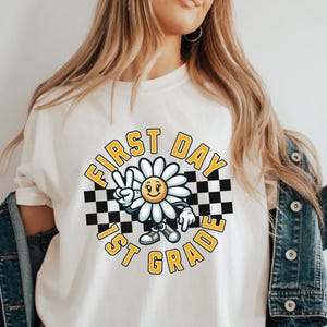 May include: White t-shirt with a graphic of a smiling daisy wearing a checkered headband and holding up two fingers in a peace sign. The graphic is surrounded by a checkered circle with the text "FIRST DAY 1ST GRADE" in yellow.