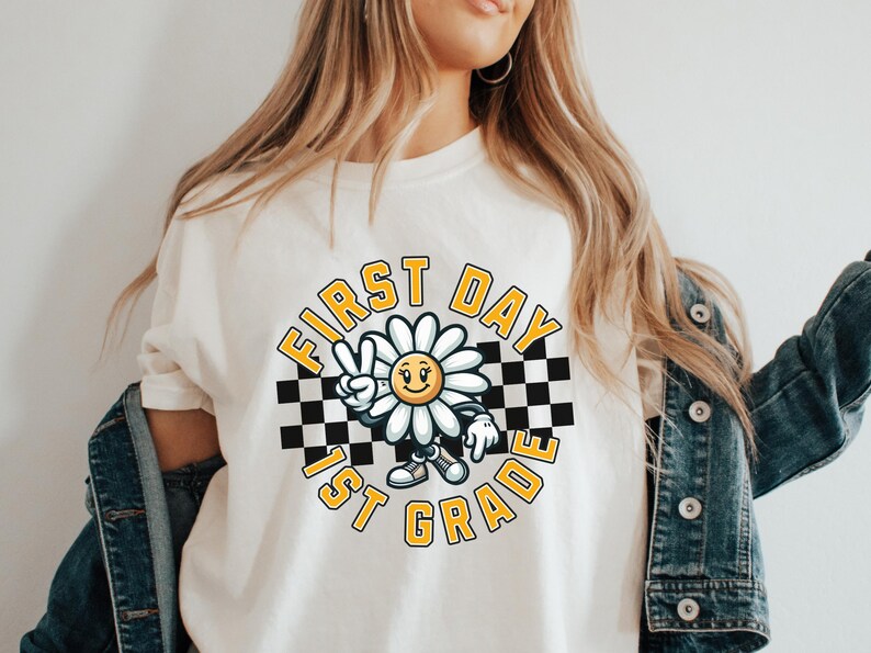 May include: White t-shirt with a graphic of a smiling daisy wearing a checkered headband and holding up two fingers in a peace sign. The graphic is surrounded by a checkered circle with the text "FIRST DAY 1ST GRADE" in yellow.