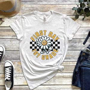 May include: White t-shirt with a graphic of a smiling daisy wearing a checkered headband and the text "First Day 1st Grade".