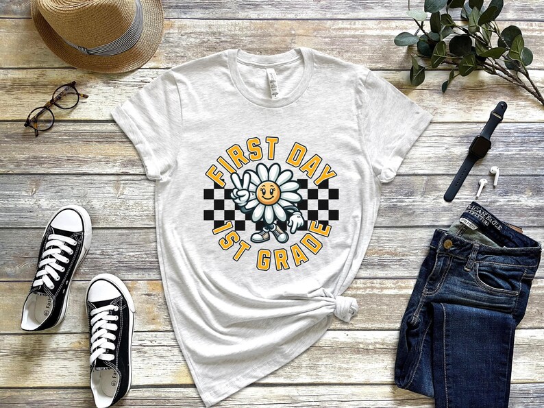 May include: White t-shirt with a graphic of a smiling daisy wearing a checkered headband and the text "First Day 1st Grade".
