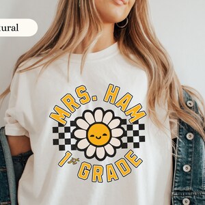 May include: Cream-colored t-shirt featuring a daisy design with a smiling face and the text "MRS. HAM 1st GRADE" in yellow and black. The daisy is centered on the shirt, with a checkered pattern on either side.