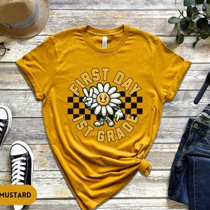 May include: A yellow t-shirt with a graphic of a smiling daisy wearing a checkered hat and the text "First Day 1st Grade".