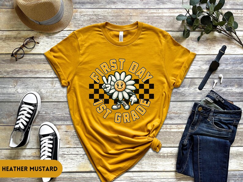 May include: A yellow t-shirt with a graphic of a smiling daisy wearing a checkered hat and the text "First Day 1st Grade".