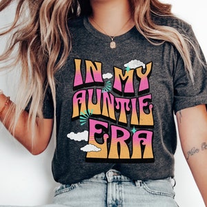 May include: Dark gray t-shirt with the text "IN MY AUNTIE ERA" in a retro font. The text is in pink, orange, and yellow, with cloud and star accents. A casual, comfortable tee.
