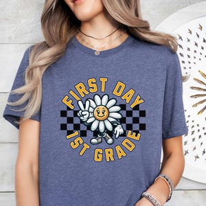 May include: A blue t-shirt with a graphic of a smiling daisy wearing a peace sign and holding a checkered flag. The text "First Day 1st Grade" is written in a yellow circle around the daisy.