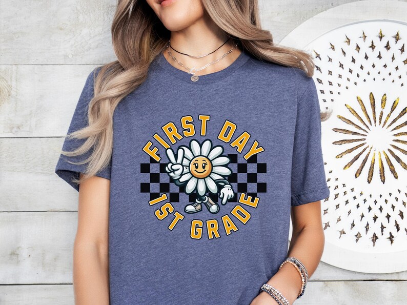 May include: A blue t-shirt with a graphic of a smiling daisy wearing a peace sign and holding a checkered flag. The text "First Day 1st Grade" is written in a yellow circle around the daisy.