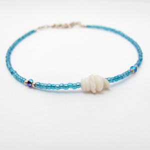 May include: A delicate blue beaded bracelet with a white seashell accent. The bracelet features small, turquoise-colored beads strung together with a silver clasp. A white, spiral-shaped seashell is the focal point of the bracelet.