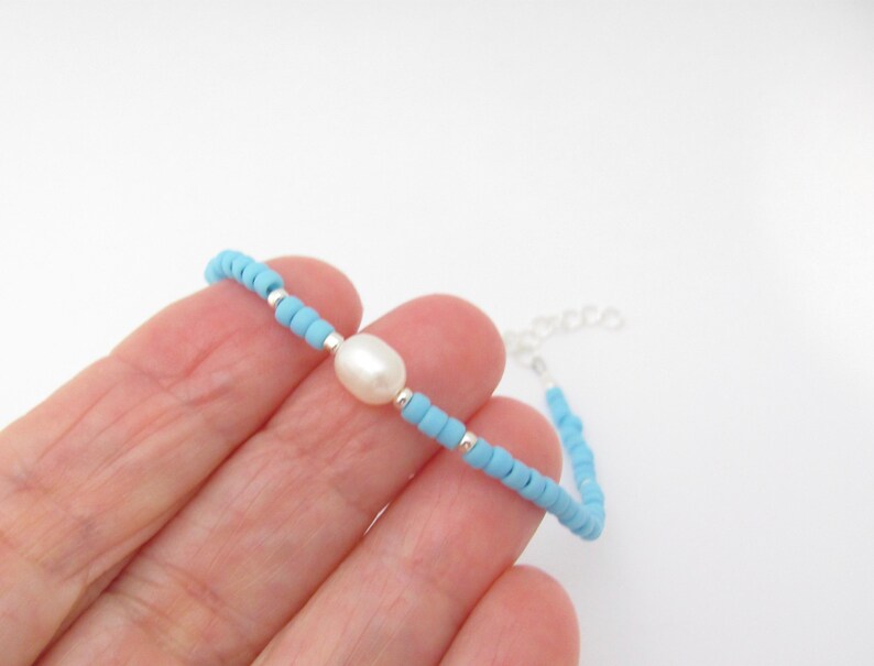 May include: A delicate bracelet made with tiny light blue beads and a single white pearl. The bracelet is secured with a silver chain.