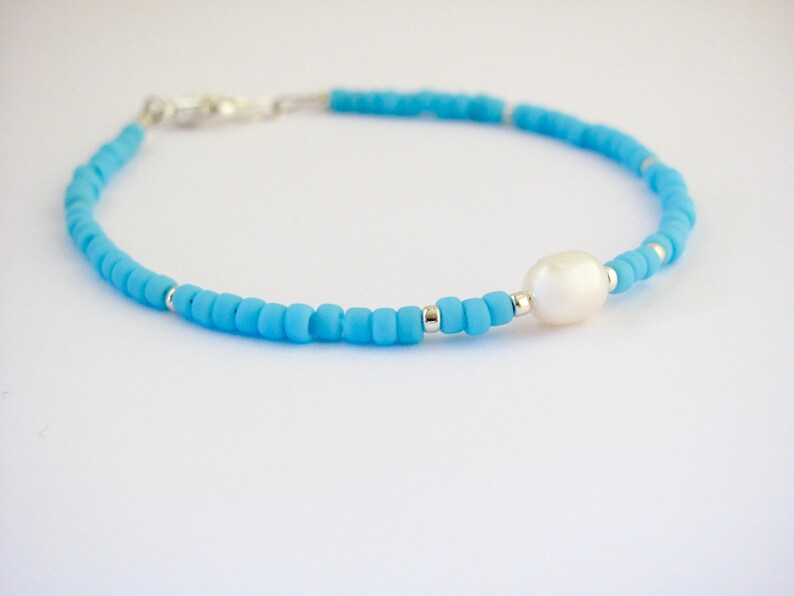 May include: A delicate blue beaded bracelet with a single white pearl accent. The bracelet is made with small, round beads and has a silver clasp.