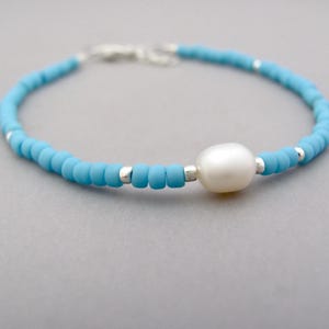 May include: A dainty bracelet featuring tiny turquoise blue beads and a solitary white pearl. The bracelet is strung on a silver chain.