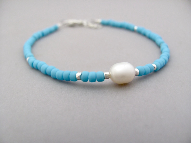 May include: A dainty bracelet featuring tiny turquoise blue beads and a solitary white pearl. The bracelet is strung on a silver chain.