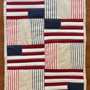 May include: A rectangular quilted table runner with a patriotic theme. The design features alternating blocks of red and white striped fabric, solid beige fabric, and navy blue rectangles, resembling the American flag. The runner is bordered with a beige fabric.
