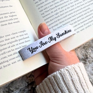 May include: A white and gray bookmark with the text "You Are My Sunshine" in black script. The bookmark is held over an open book, with the text of the book visible in the background. The bookmark is a simple, yet elegant design.