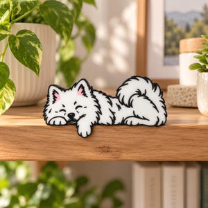 May include: A white, flat, decorative dog figurine with black outlines, depicting a fluffy canine lying down with its head resting on its front paws. The dog has pointed ears with pink inner details and a curled, bushy tail.