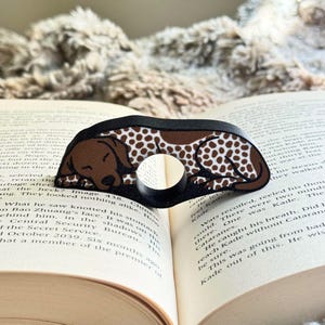 May include: A brown and white dog-shaped book page holder rests on an open book. The page holder has a circular opening and a black border. The dog design features a sleeping dog with brown spots.