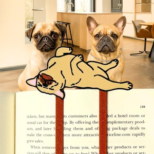 May include: A bookmark featuring a cartoon French Bulldog lying on its back, placed in a book. Two real dogs, a Pug and a French Bulldog, are in the background. The book has text on the pages and a red ribbon.