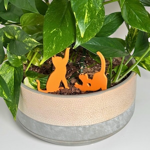 May include: Two orange cat silhouettes are used as plant stakes in a pot of green leafy plants. The pot is a light brown color with a gray band around the top.