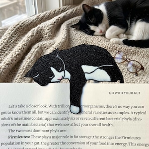 May include: A black and white cat-shaped bookmark resting on a book, with the text "GO WITH YOUR GUT" visible. A sleeping cat is in the background. The bookmark has a pink nose and closed eyes.