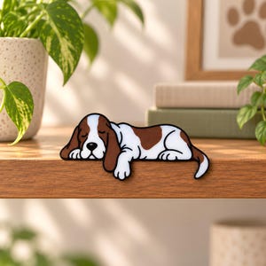 May include: A flat, decorative cutout of a sleeping basset hound dog with white and brown fur patches. The dog is depicted in a resting position with its head tucked down and eyes closed, featuring a black outline and a black nose.