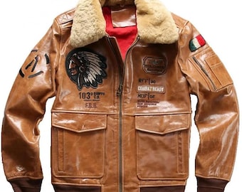 Puede incluir: Una chaqueta bomber de cuero marrón con cuello de piel sintética. La chaqueta tiene varios parches, incluyendo una cabeza de indio americano, un parche militar y un parche con el texto "Combat Ready".