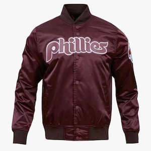 Puede incluir: Una chaqueta de béisbol de satén marrón con la palabra "Phillies" en letras blancas en la parte delantera. La chaqueta tiene una cremallera y un cuello, puños y dobladillo de canalé.