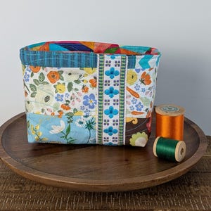 May include: A patchwork fabric basket with floral and geometric patterns. The basket features a blue trim and a decorative ribbon with blue floral embroidery. Two spools of thread, one orange and one green, sit beside the basket on a wooden tray.