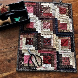 May include: A small quilt with a log cabin pattern in shades of red, brown, and white. The quilt is made of various fabrics with different patterns and textures. A pair of scissors is lying on top of the quilt.