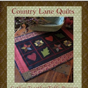 May include: A dark blue table runner with a red and white border. The runner features a patchwork design with hearts, stars, and flowers. The text "Gather Together Table Runner" and "by Kathleen Tracy" is printed below the runner. The website "www.countrylanequilts.com" is printed below the text.