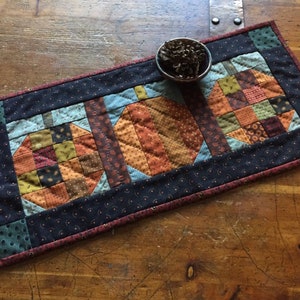 May include: A quilted table runner featuring three patchwork pumpkins in shades of orange, brown, and green. The pumpkins are set against a blue background with a dark border. A small metal bowl with pinecones sits on the runner.