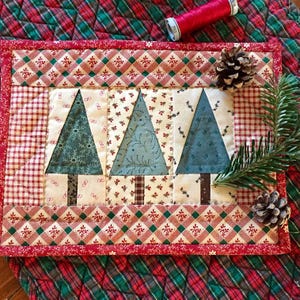 May include: A quilted Christmas placemat featuring three teal Christmas trees with brown trunks. The placemat has a red and white checkered border and a red floral patterned border. A pine cone and sprig of pine are on the right side.