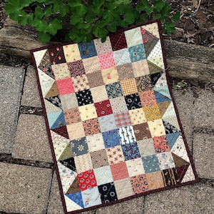 May include: A patchwork quilt with a variety of colorful fabrics in a square pattern. The quilt is made up of small squares of different colors and patterns, including red, blue, green, and white. The quilt is edged with a dark red border.