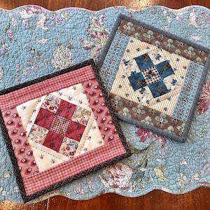 Op de afbeelding: Twee vierkante, gewatteerde pannenlappen met ingewikkelde patchworkontwerpen. Eén heeft rode en witte bloemen- en ruitpatronen, de andere blauwe en bruine bloemen- en streepstoffen. Ze liggen op een lichtblauwe bloemen gewatteerde placemat.