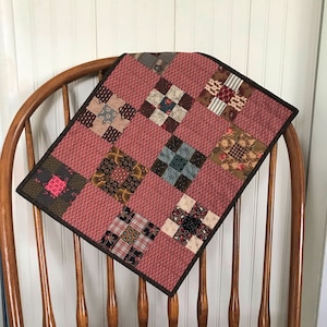 May include: A handmade quilt with a patchwork design. The quilt features a red background with square blocks of various patterned fabrics in shades of brown, blue, and cream. The quilt is trimmed with a dark brown border and is draped over a wooden chair.