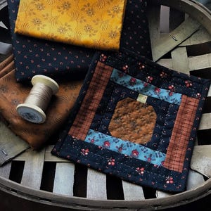 May include: A small quilt block with a pumpkin design in shades of brown, blue, and black. The quilt block is made with different fabrics and is surrounded by a basket weave pattern. A spool of thread is also visible in the image.