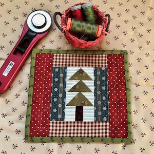 May include: A handmade Christmas tree quilt block with a red rotary cutter and a small basket of thread spools. The quilt block features a tree design with green, blue, and brown fabrics, and is bordered by red and green fabrics.