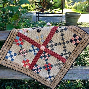 May include: A patchwork quilt with a red, white, and blue color scheme. The quilt is made of squares of different fabrics and is sewn together in a pattern. The quilt is displayed on a wooden fence.