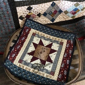 May include: A small quilt with a star pattern in shades of brown, red, and cream, surrounded by a border of blue and white fabric. The quilt is sitting in a wooden basket with a pair of scissors on top.