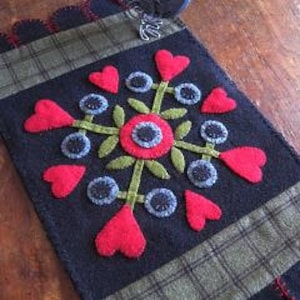 May include: A black felt rug with a green and red floral design. The design features a central flower with eight surrounding hearts and leaves.
