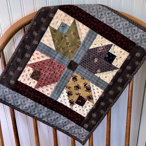 May include: A quilt block with a patchwork design featuring various floral patterns in shades of brown, green, and white. The block is framed with a dark brown border and a light gray border with a small pattern.
