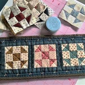May include: A blue and brown quilted table runner with three squares featuring different patchwork patterns. The squares are surrounded by a blue border with a brown outer border. The text "Eat Sleep Quilt" is on a blue tin container.