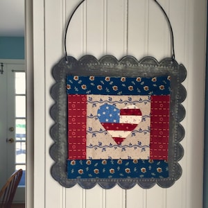 May include: A patriotic heart-shaped quilt block with red, white, and blue fabric, framed in a distressed metal frame with a scalloped edge. The quilt block is hanging on a white wall.