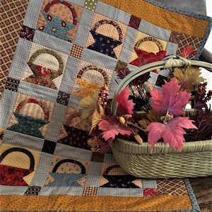 May include: A patchwork quilt with a blue and white checkered background and a pattern of brown, red, and yellow baskets. The quilt is folded and placed on a wooden surface with a woven basket filled with fall leaves.