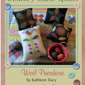 May include: Wool pincushions in various shapes and colors, including a flower, a star, and a circle. The pincushions are displayed on a patchwork quilt with buttons and a basket of yarn. The text "Wool Pincushions by Kathleen Tracy www.countrylanequilts.com" is displayed below the pincushions.