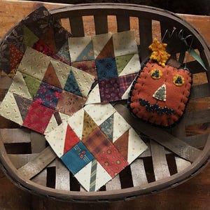 May include: A wooden basket holds several patchwork quilt squares and a felt pumpkin decoration. The quilt squares feature various patterned fabrics in shades of red, blue, green, and beige. The pumpkin is orange with felt facial features.