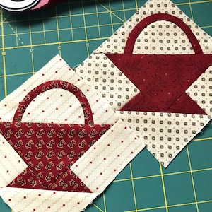 May include: Two red and white fabric patchwork squares with a basket design. The baskets are made of red fabric with a white floral pattern and have a white fabric handle.
