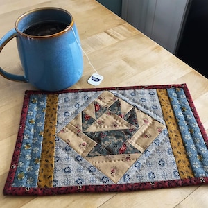 May include: A blue ceramic mug filled with tea and a tea bag rests on a handmade quilted placemat. The placemat features a central geometric design in beige and floral patterns, with blue and gold side panels.