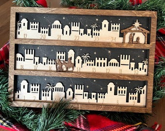 May include: A wooden advent calendar with a Christmas village scene. The calendar has three rows of houses and a star on top. The numbers 1 through 24 are visible on the calendar.