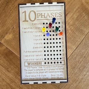 May include: Wooden board game with the text "10 PHASES" and colorful pegs. The game board has a series of rules and instructions printed on it, including "WINNER!" and point values. The game is designed for 2-6 players.
